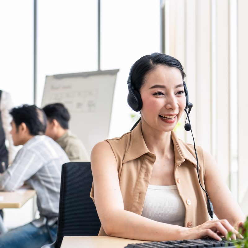 Asian young beautiful call center work with smile in workplace while colleague meeting and brainstorming to solve problem and discuss in workplace background. Business Network, Communication concept.