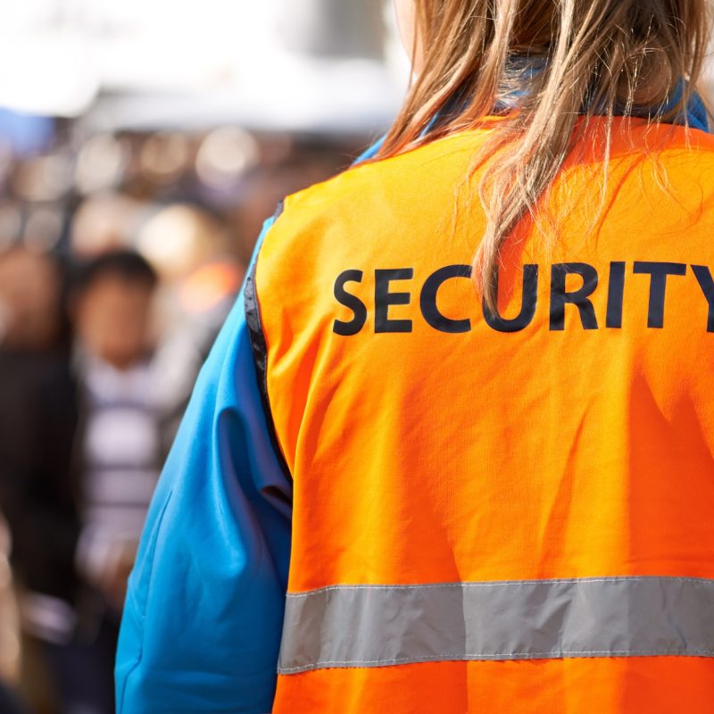 Rearview shot of a security officer standing outdoors with a crowd in the background.