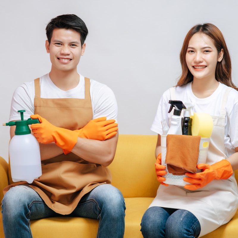 happy smiling young couple wearing gloves and holding equipment and spray bottle for cleaning the house together in living room